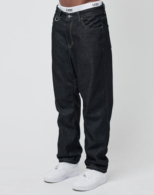 Someone wears loose-fitting Work Denim jeans by LIVE FAST DIE YOUNG, white sneakers, and shows the waistband of white LFDY underwear. The deep indigo denim stands out against a plain, light backdrop.