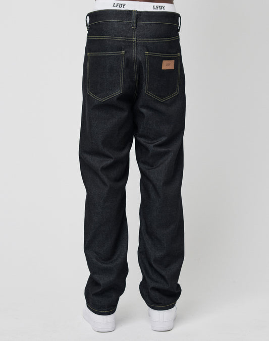 A person shown from the back wears LIVE FAST DIE YOUNG’s Work Denim jeans in dark blue with yellow stitching, styled with white sneakers and a logo waistband, against a plain white background.