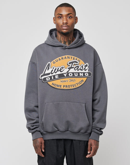 A man with short hair and neck tattoos models the LIVE FAST DIE YOUNG Home Protection Hooded, featuring an oversize fit and bold front print, standing against a plain light gray background.