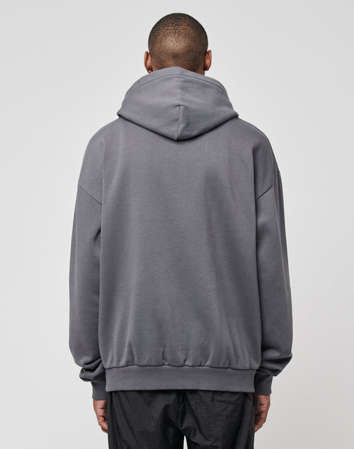 A person faces away from the camera, wearing the LIVE FAST DIE YOUNG Home Protection Hooded in an oversize fit gray style with black pants, set against a plain light gray background.