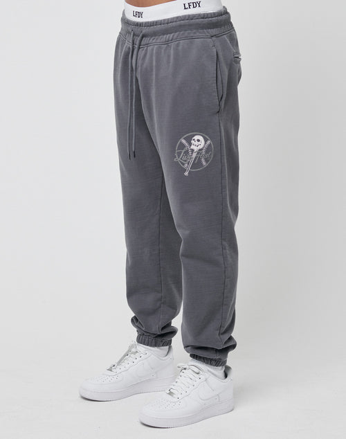 Model wears the LIVE FAST DIE YOUNG Live Fast Skull Sweat Jogger—gray cotton joggers with a white skull and lollipop graphic on the left thigh, branded white LFDY waistband, and white sneakers, shown in a regular fit against a plain background.