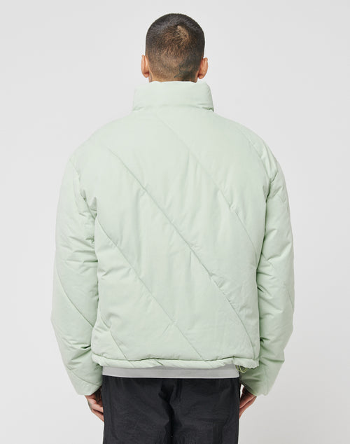 A person with short hair is seen from behind in a light green LFDY Puffer Jacket with an oversize fit, diagonal stitching, and a high collar, paired with black pants. The plain white background highlights the jacket’s design.