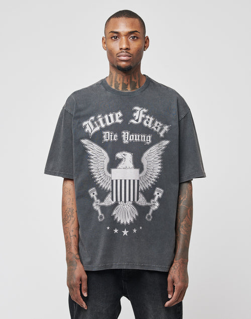 A man wears the LIVE FAST DIE YOUNG Eagle Tee, a faded black 100% cotton regular fit with a bold eagle front print, shield, and “Live Fast Die Young” text, standing against a plain light background.