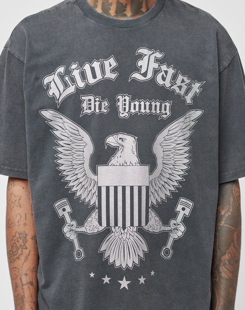 A person wears the Eagle Tee by LIVE FAST DIE YOUNG, a dark regular fit T-shirt with a bold eagle holding pistons and a shield on the front, Live Fast Die Young text above, and tattoos visible on their neck and arms.