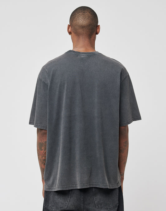 A person with short hair stands with their back to the camera, wearing the LIVE FAST DIE YOUNG Eagle Tee in faded black (100% Baumwolle, Regular Fit) and black jeans. Their tattooed arms are visible against a plain light gray background.