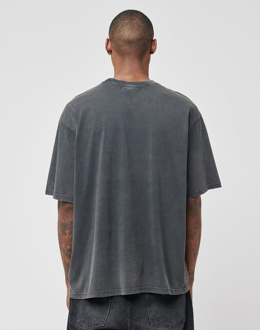 A person with short hair stands with their back to the camera, wearing the LIVE FAST DIE YOUNG Eagle Tee in faded black (100% Baumwolle, Regular Fit) and black jeans. Their tattooed arms are visible against a plain light gray background.