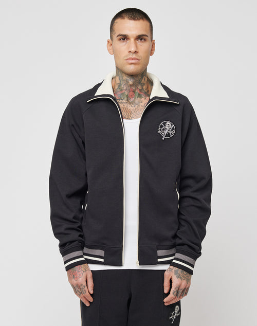 A man with short hair and visible neck and chest tattoos wears a black LIVE FAST DIE YOUNG Track Suit Jacket with white trim over a white tank top. The Regular Fit jacket, made from Polyester Baumwolle, has a small white graphic on the left chest.