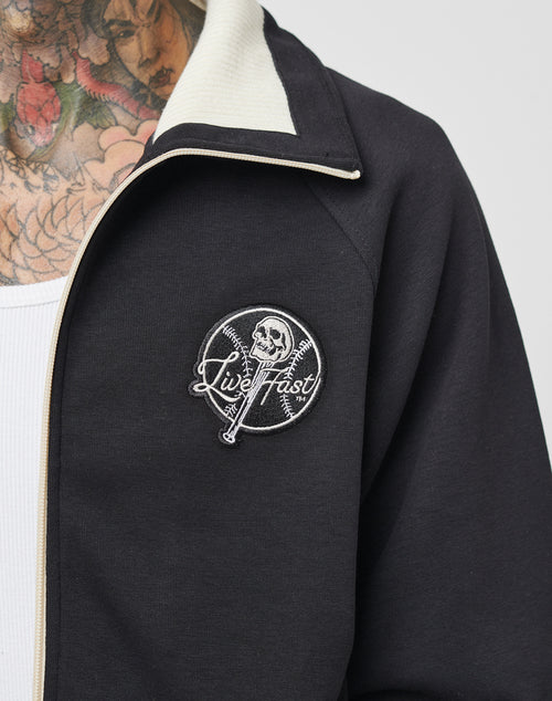 A close-up of a person wearing a dark LIVE FAST DIE YOUNG Track Suit Jacket with a white collar and “Live Fast” patch showing a skull and crossed bats. Their neck and chest feature colorful tattoos.