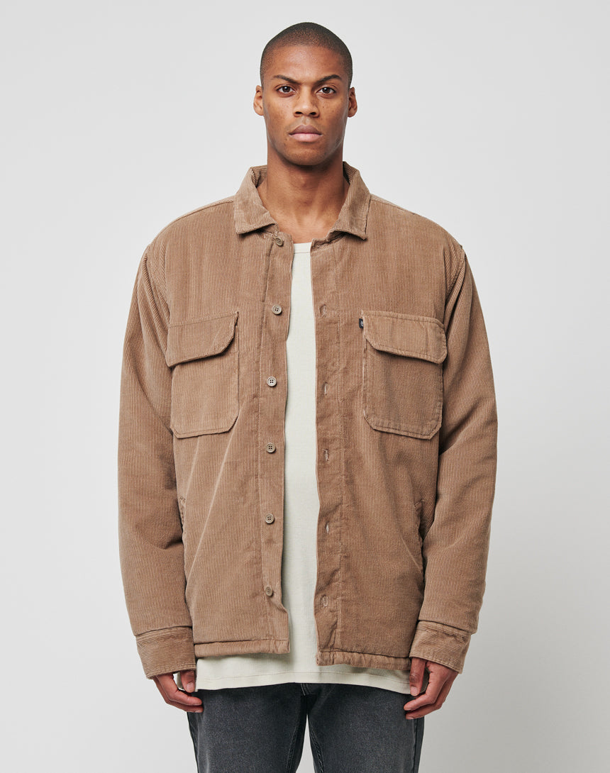 A person faces forward in a light brown LFDY Corduroy Shacket with regular fit, worn over a cream shirt and dark pants. The shacket features a quilted lining. The background is plain and light-colored.