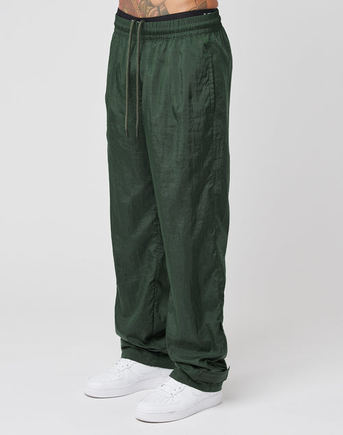 A person wears LIVE FAST DIE YOUNG Nylon Jogger pants in dark green with a crinkled effect and elastic waistband, styled with white sneakers against a plain white background.