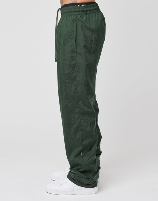 A person in LIVE FAST DIE YOUNG Nylon Jogger—dark green, loose-fit with wide elastic waistband, drawstring, and subtle Flag Label—pairs them with white sneakers while standing in profile against a plain white background.