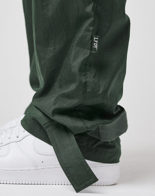 A close-up of a person wearing LIVE FAST DIE YOUNG Nylon Jogger in dark green with a wide elastic waistband, adjustable ankle straps, LFDY branding above the strap, white sneakers, and a subtle flag label for detail.