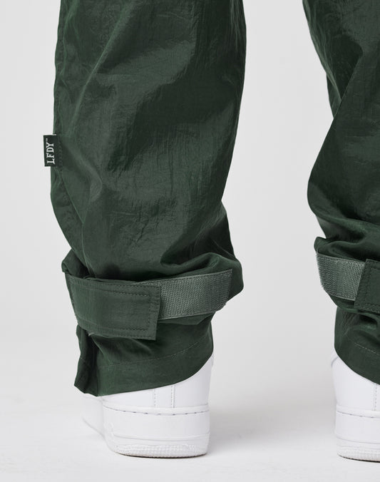A close-up of the lower legs in LIVE FAST DIE YOUNG Nylon Jogger pants with Crushed Effekt, adjustable cuff straps, and a Flag Label, paired with white sneakers on a white background.