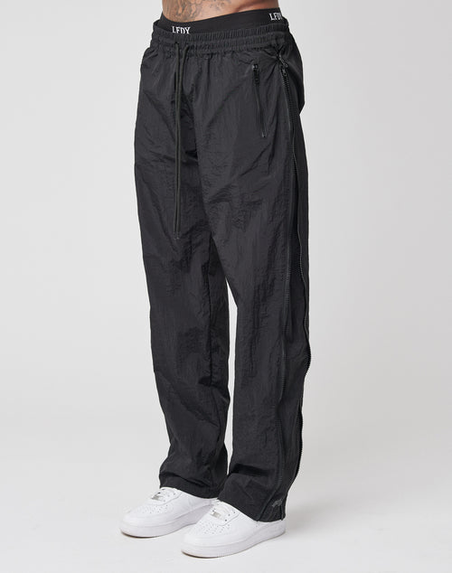 A person wearing LFDY Zip Tape Track Pants in black nylon with a crinkled texture and zippered pockets, paired with white sneakers, stands against a plain white background. Only the lower body is visible.