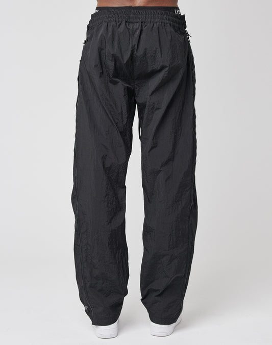 A person wearing LFDY Zip Tape Track Pants in black nylon with a crinkled texture and elastic waistband, viewed from the back. The regular fit pants are paired with white shoes against a plain white background.