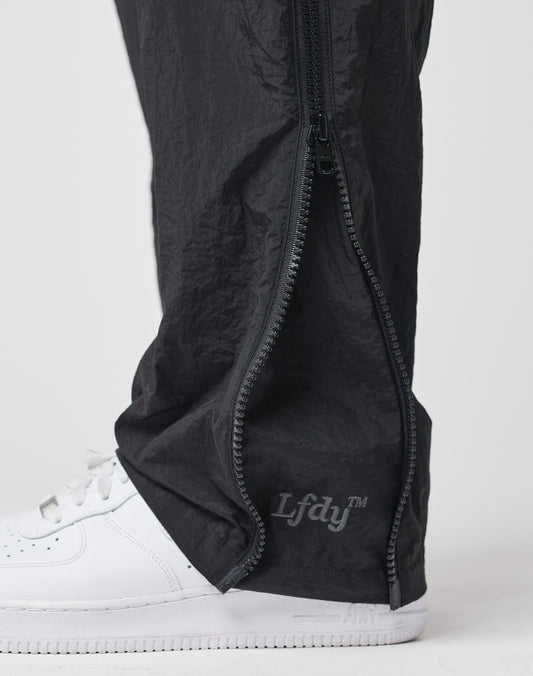 Close-up of a person wearing LFDY Zip Tape Track Pants in black nylon, with the ankle zipper partially open to show a white sneaker and the LFDY logo near the ankle.