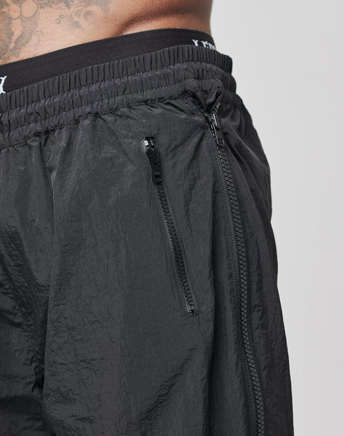 Close-up of a persons waist wearing LFDY Zip Tape Track Pants in black nylon with a crinkled texture, elastic waistband, and visible zippers on the fly and side pocket; part of their tattooed torso is also visible.