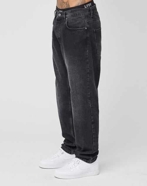 The lower half of a person wearing LIVE FAST DIE YOUNG Regular Denim—regular fit, faded black jeans aus Baumwolle with a subtle Lederpatch—and white sneakers is visible against a plain light gray background.