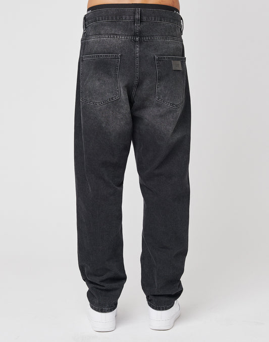 A person is shown from the back wearing LIVE FAST DIE YOUNG Regular Denim jeans—loose-fitting, faded black, 100% Baumwolle—with two large back pockets, visible stitching, a subtle Lederpatch, and white sneakers against a plain background.