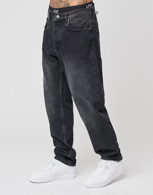 A person wears loose-fitting, faded black Regular Denim jeans by LIVE FAST DIE YOUNG, with cuffed hems and LFDFV text on the waistband, paired with white sneakers against a plain white background.