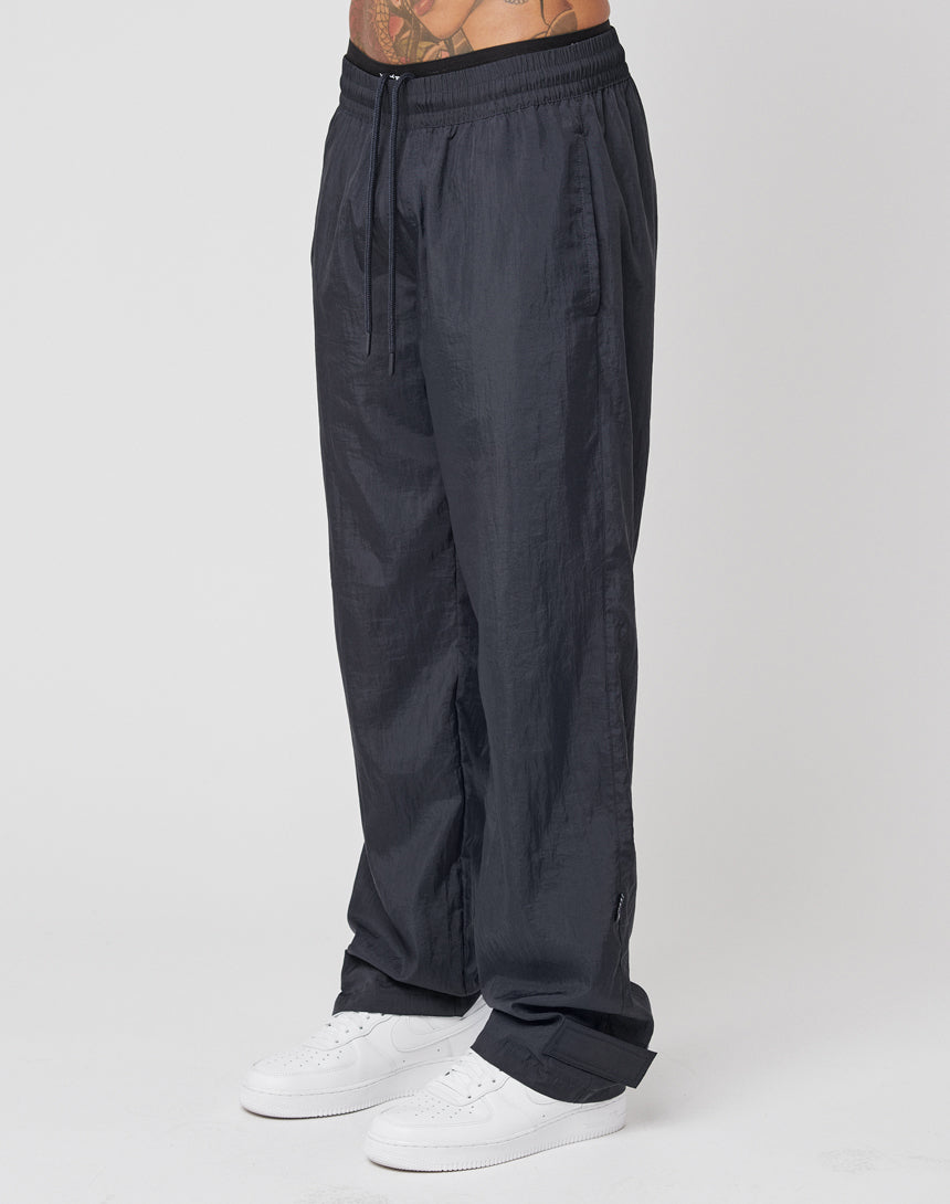 A person wears LIVE FAST DIE YOUNG Nylon Jogger track pants in dark gray with a wide elastic waistband and drawstring, paired with white sneakers. The photo is cropped from lower torso to feet.