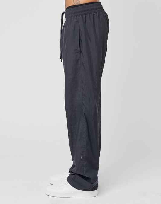 A person in profile wears LIVE FAST DIE YOUNG Nylon Jogger—regular fit, black nylon pants with a wide elastic waistband and drawstring—paired with white sneakers; a tattoo is visible on their lower torso against a plain white background.