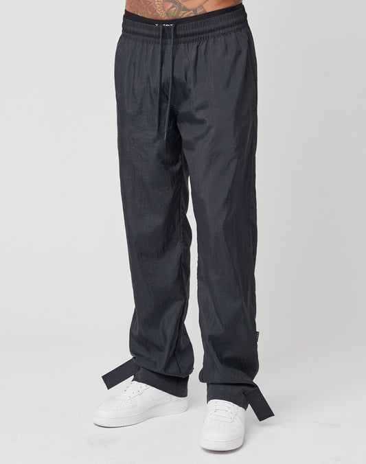 A person is shown from the waist down wearing LIVE FAST DIE YOUNG Nylon Jogger pants in black with a wide elastic waistband, drawstring, ankle straps, and white sneakers against a plain white background.