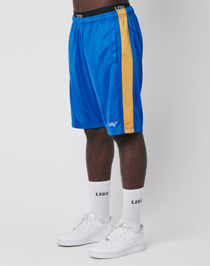 A person wears LFDY Taped Stripe Shorts—blue with a yellow side stripe and elastic waistband—made from polyester, paired with white crew socks featuring LFDY text and white sneakers, standing against a plain white background.