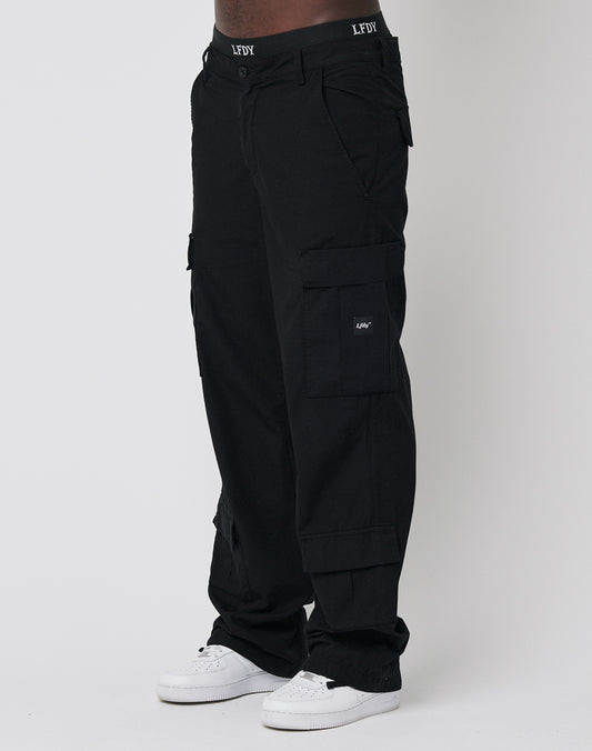 A person in wide fit black LFDY Wide Summer Cargo pants with multiple pockets and LEFY branding on the waistband and pocket, paired with white sneakers, stands against a plain light background. | model