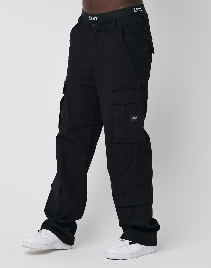 A person in black LFDY Wide Summer Cargo pants with multiple pockets and a wide fit in comfortable cotton, paired with white sneakers, stands against a plain white background. The LFDY logo appears on the waistband and pocket.