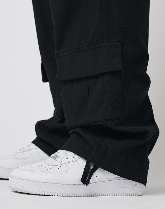 A close-up of a person wearing LFDY Wide Summer Cargo pants in black and white Nike Air Force 1 sneakers, against a plain white background. The Wide Fit cargos feature a large pocket and ankle drawstring detail.