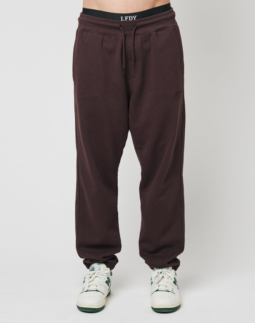 Only the lower half of a person is visible, wearing LFDY Sweat Jogger 3.0 in dark brown with elastic waistband and drawstring, paired with white sneakers, set against a plain white background.