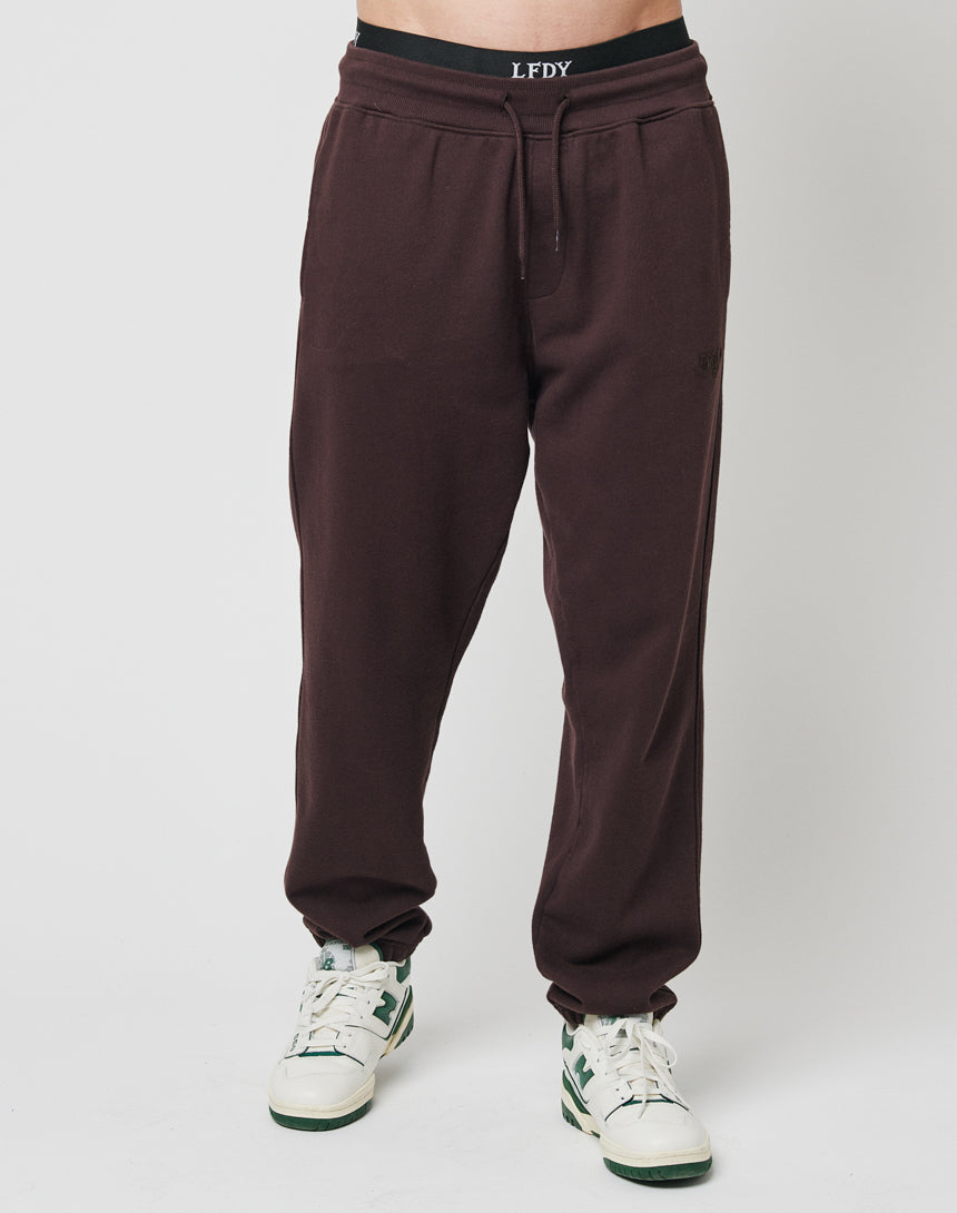 A person wearing LFDY Sweat Jogger 3.0 in dark brown with elastic waistband and white sneakers with green details, standing against a plain white background.