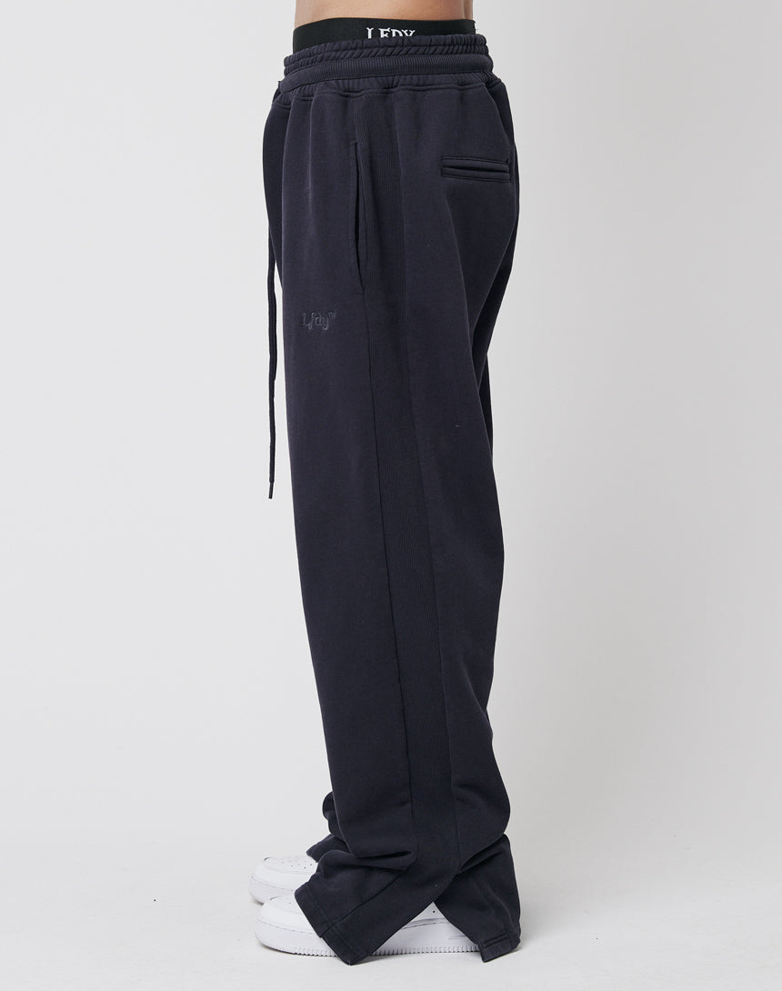 A person stands in profile against a plain white background, wearing LFDY’s Wide Jogger 2.0—wide fit cotton joggers with side and back pockets, a drawstring waist, and white sneakers.