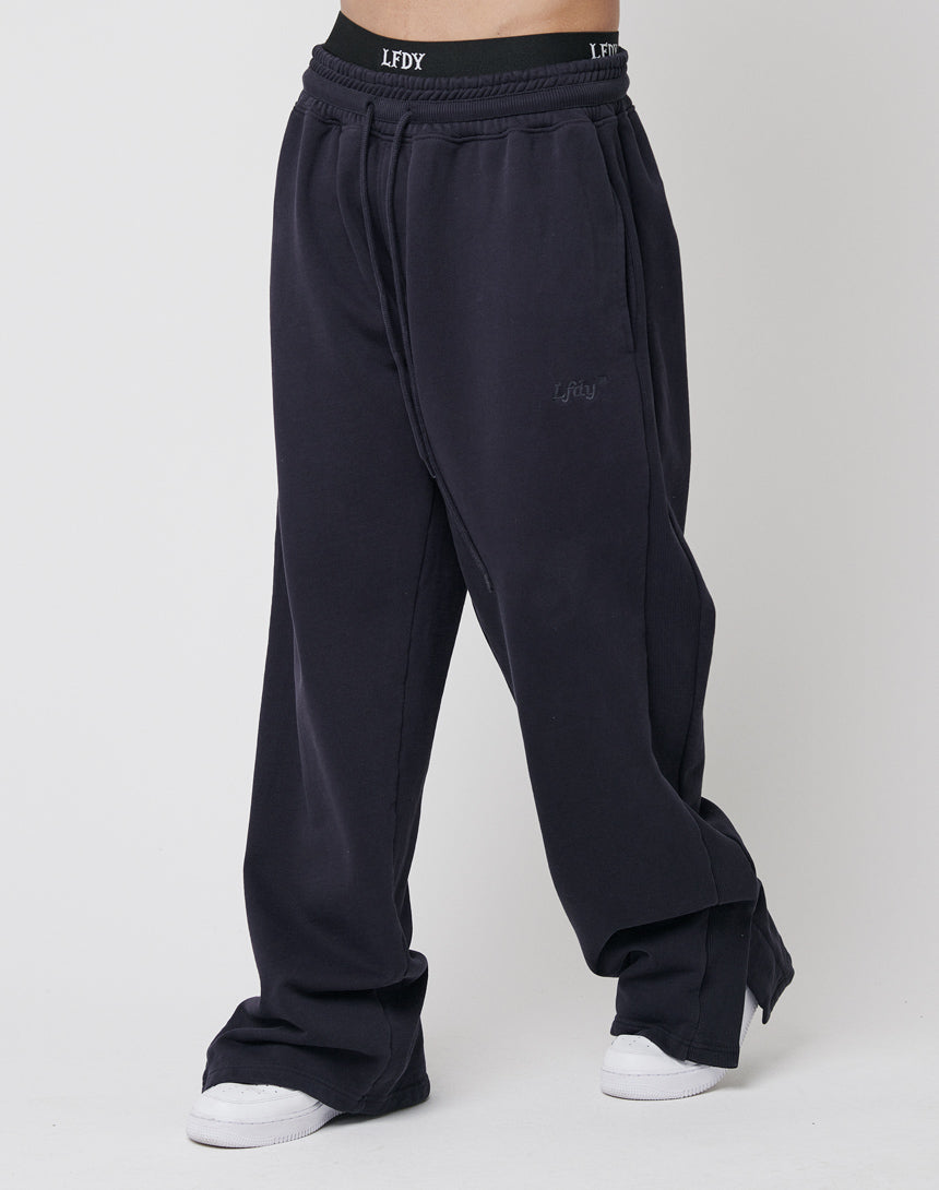 A person wears the LFDY Wide Jogger 2.0, a loose-fitting black jogger with an elastic waistband branded "LFDY," made from soft cotton, styled with white sneakers. Only the lower body is visible against a plain background.