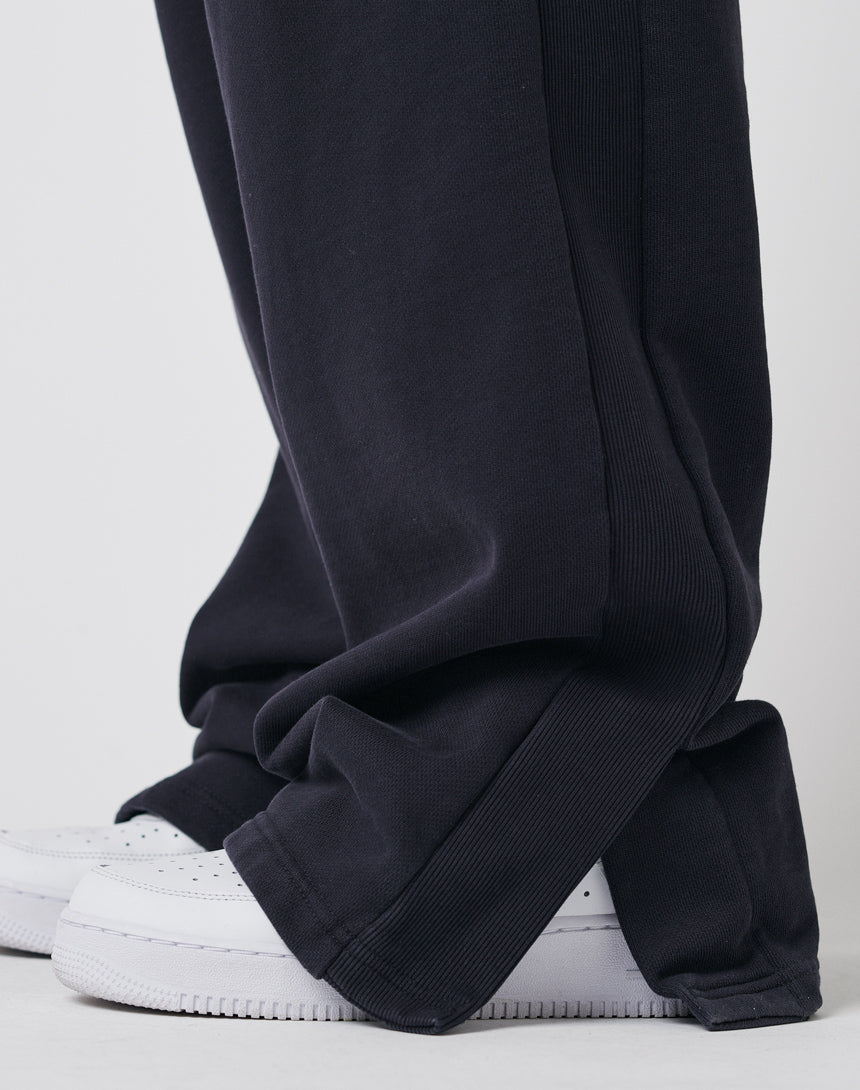 A close-up of a person wearing LFDY Wide Jogger 2.0 in dark blue, featuring wide legs that drape over white sneakers. The ribbed cotton pants bunch slightly at the ankles, partially covering the shoes.