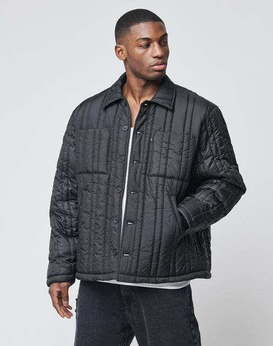 A man wears a black Import & Export Jacket by LIVE FAST DIE YOUNG over a white shirt, standing against a plain light gray background. He gazes slightly left, with one hand by his side and the other partially in his pocket.