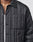 Close-up of a person wearing the Import & Export Jacket by LIVE FAST DIE YOUNG over a white shirt. The black, quilted oversize jacket has vertical stitching, a 3D chest pocket, collar, and visible buttons; only lower face and upper torso are visible.